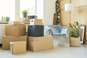 Stacks of large boxes standing against windowsill with green domestic plants in flowerpots and corner with rolled carpets and armchair
